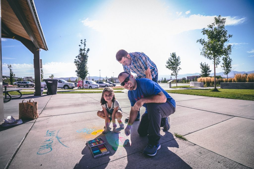 Two men and little girl kneeling down by sidewalk chalk drawing and smiling.