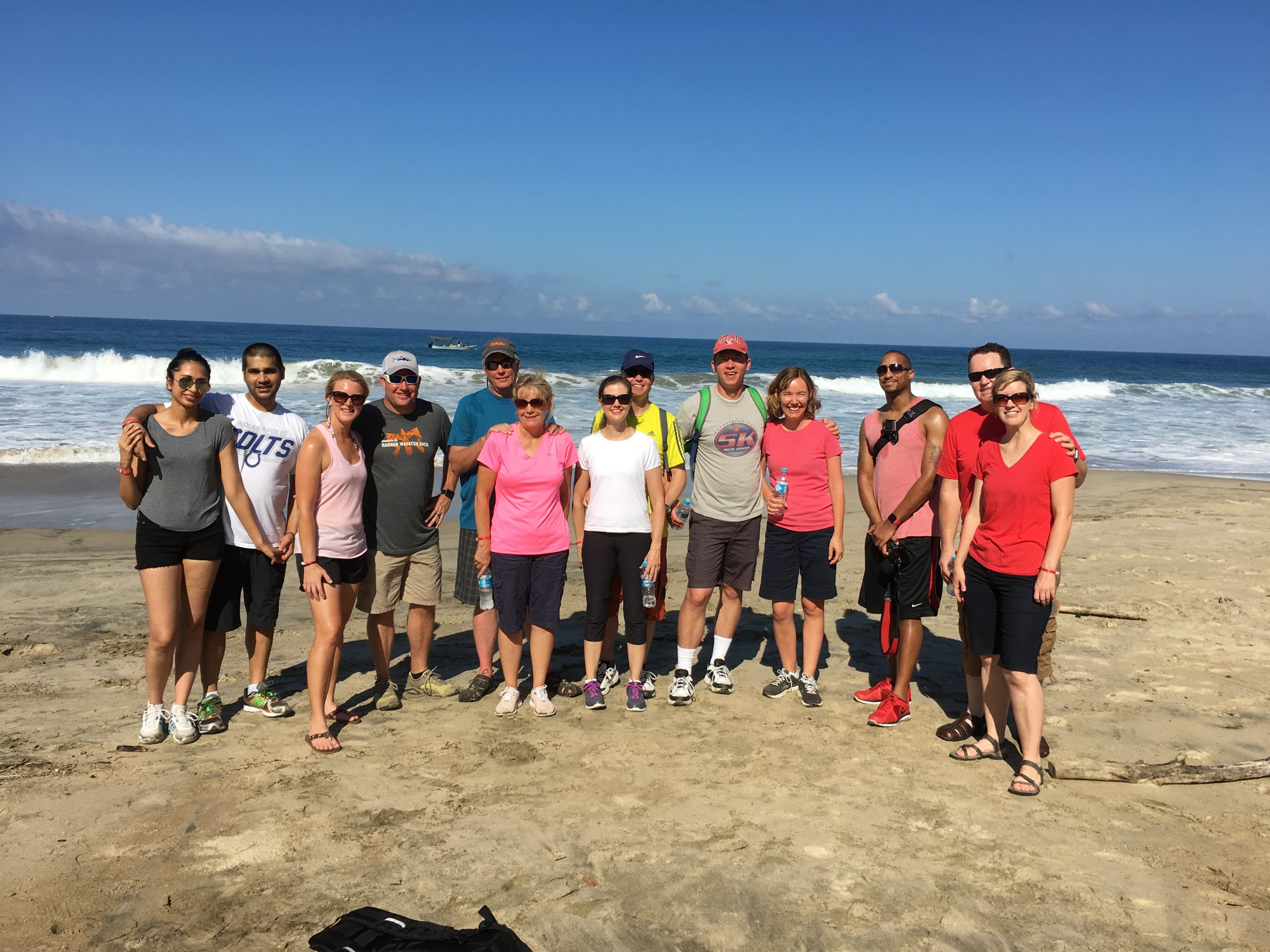 Group of men and women standing and smiling for photo on sandy beach with ocean in the background.