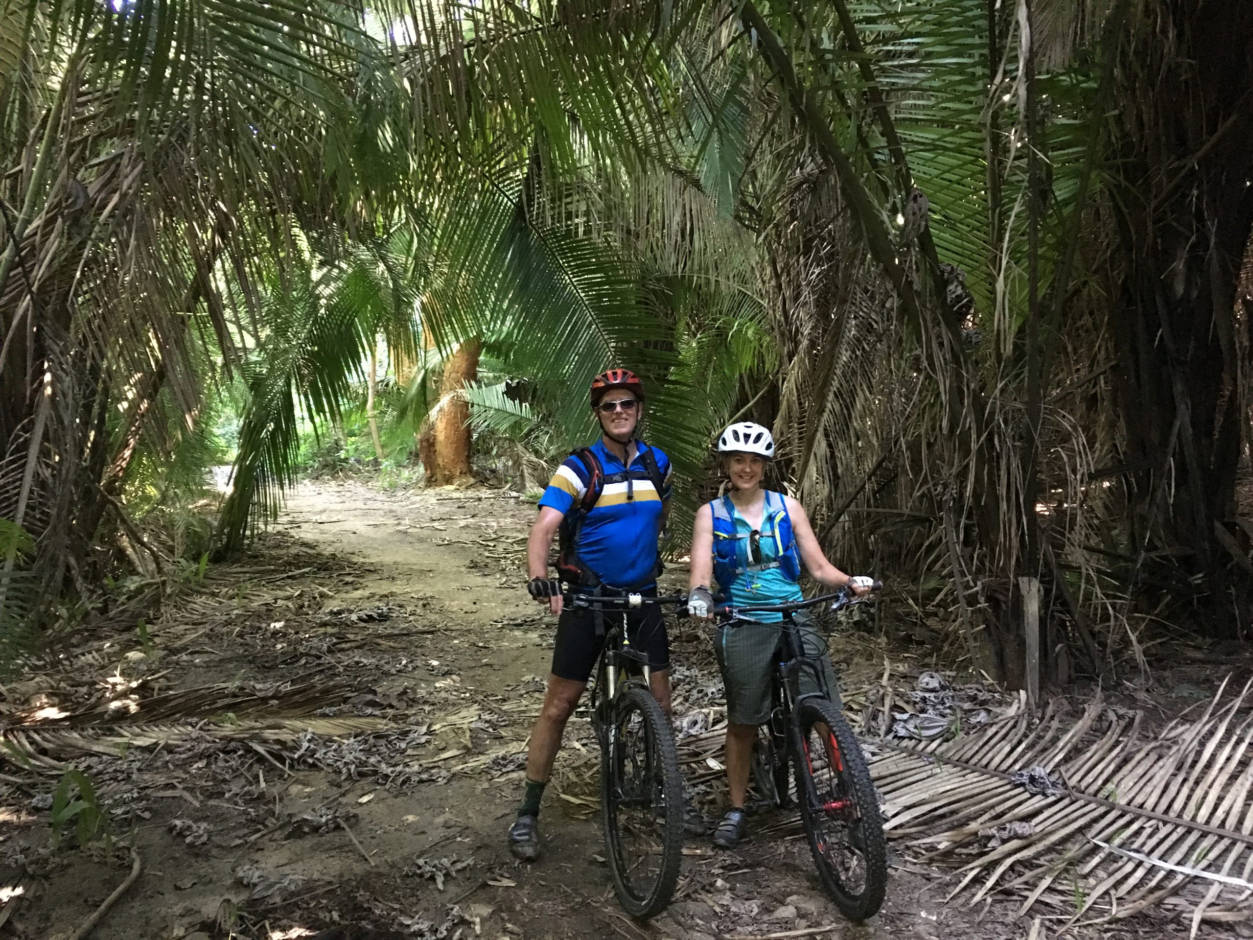 Man and woman standing with mountain bikes in helmets and mountain bike attire on dirt trail surrounded by palm trees.