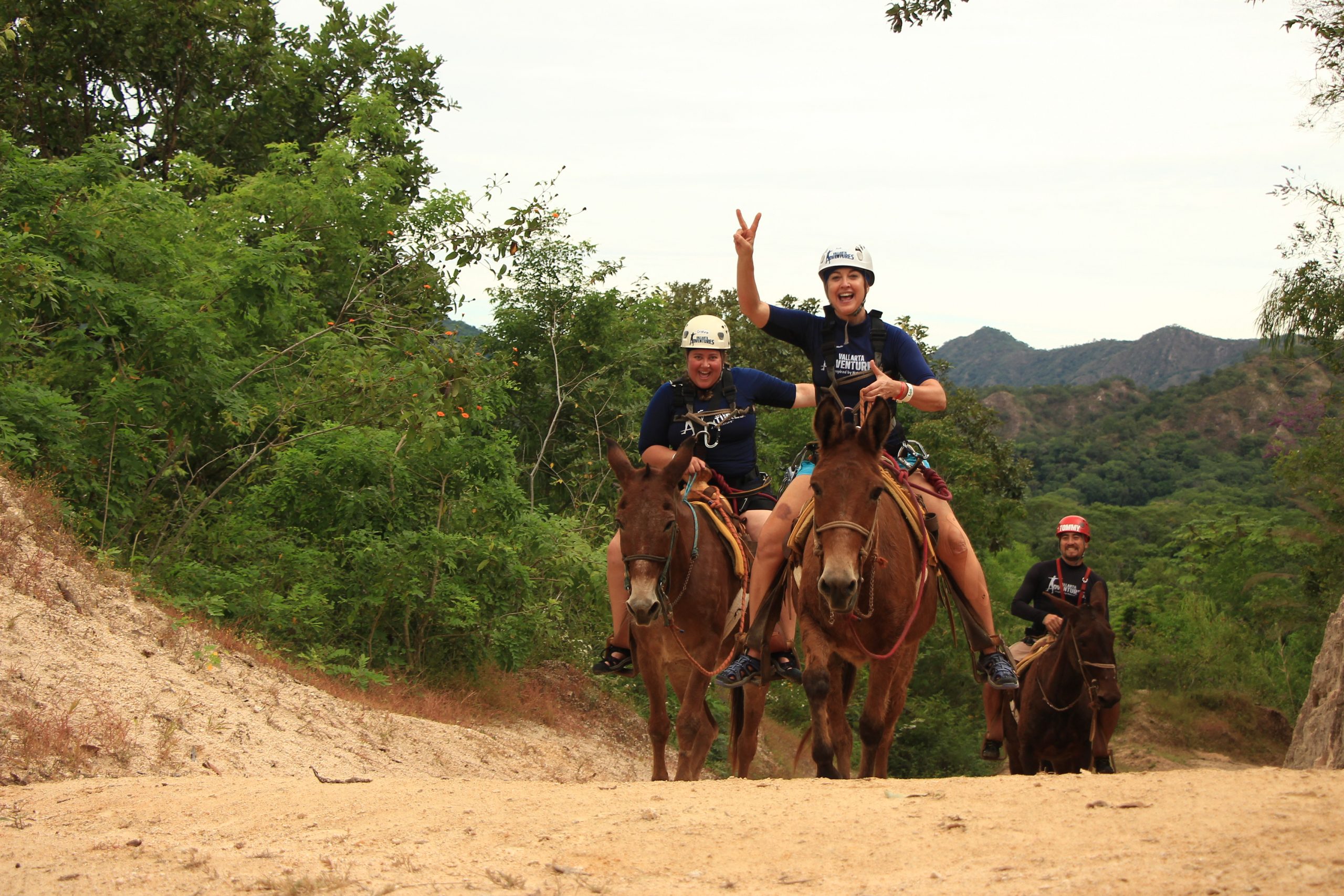 Two women and a man riding horses on dirt trail while smiling at camera.