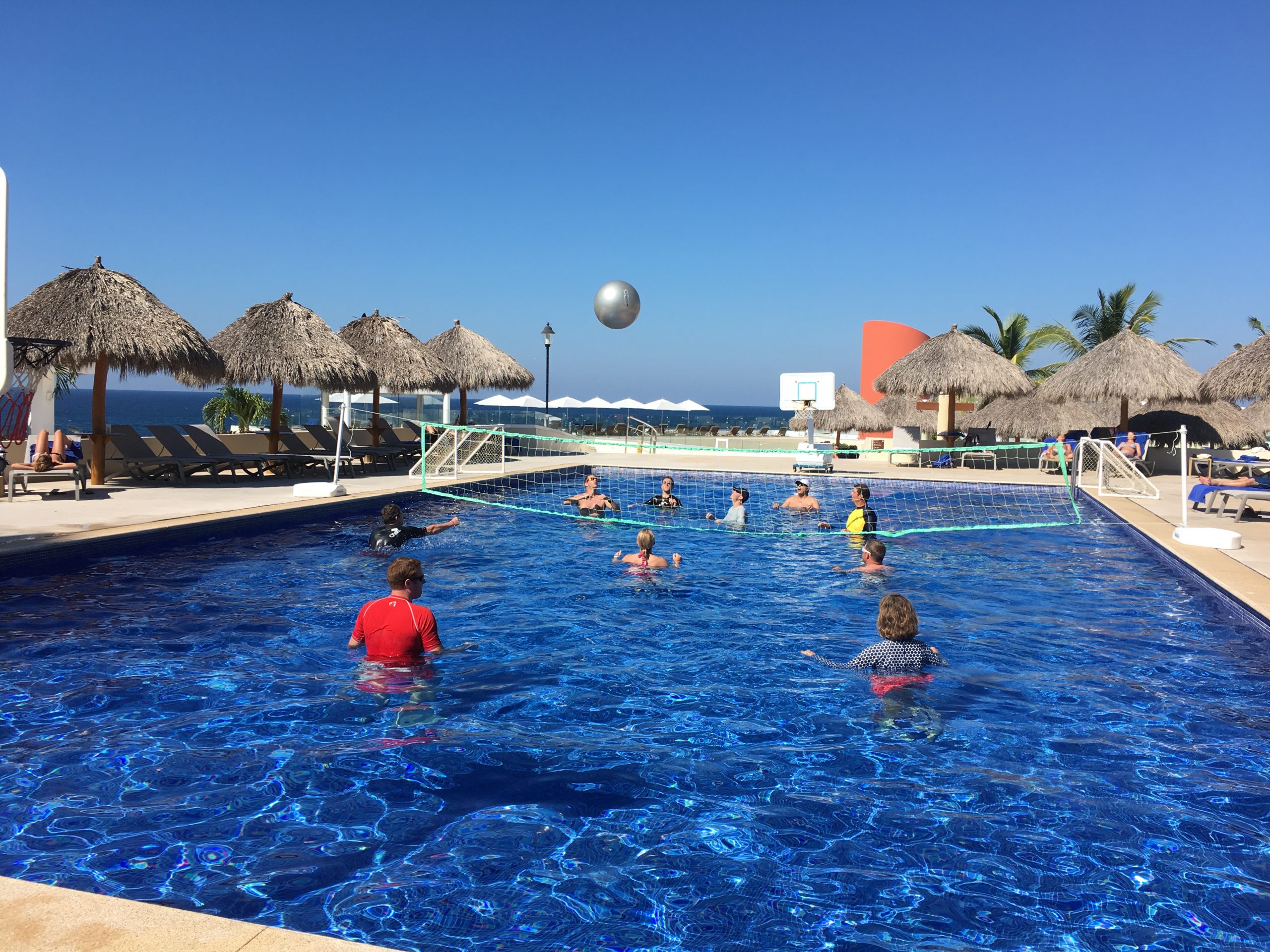 People playing volley ball in swimming pool at resort hotel with ocean view in the background.