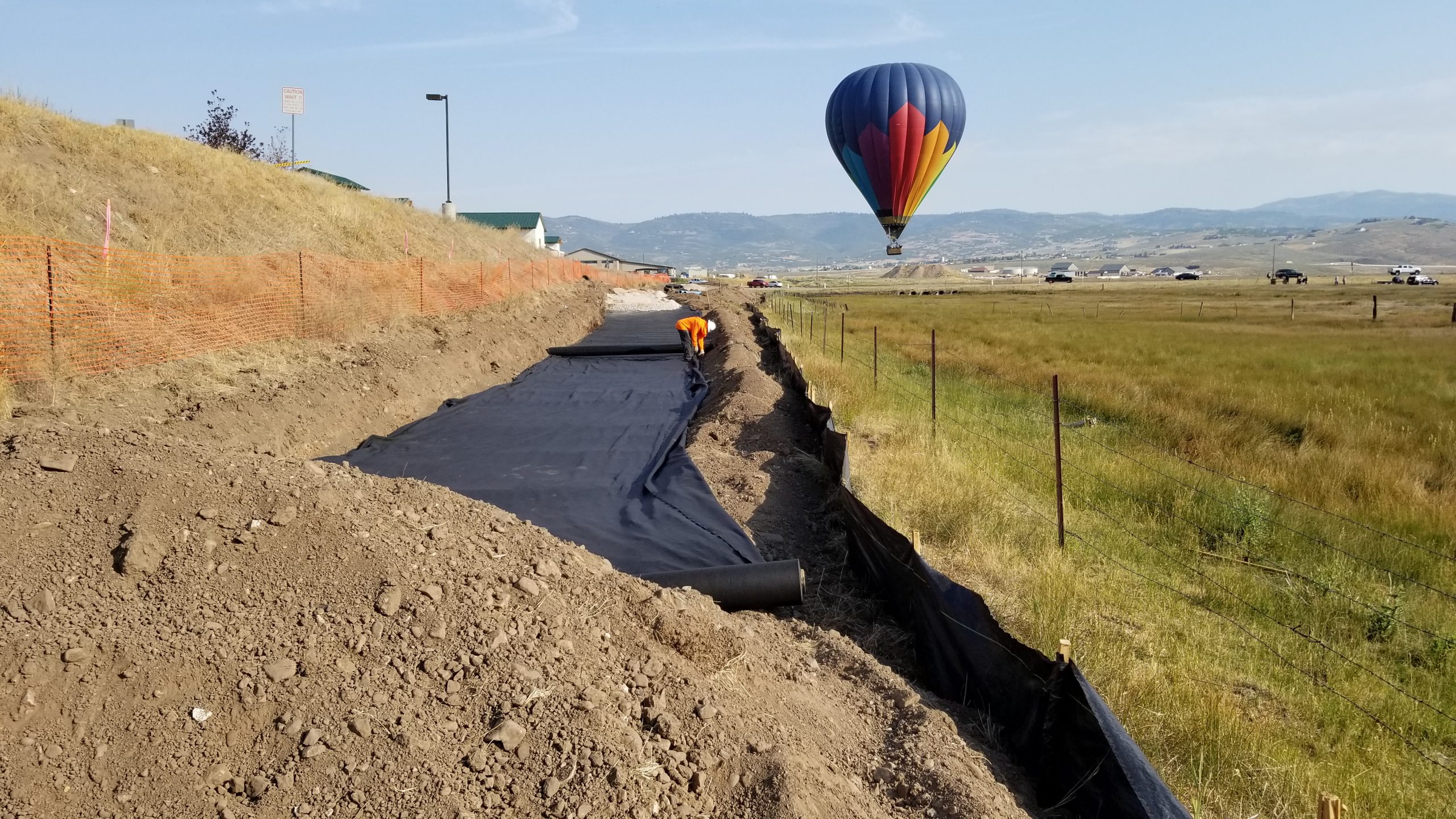 Construction worker laying down black tarp on site of Silver Creek Trunkline Sewer Rehabilitation project with hot air balloon in the background.