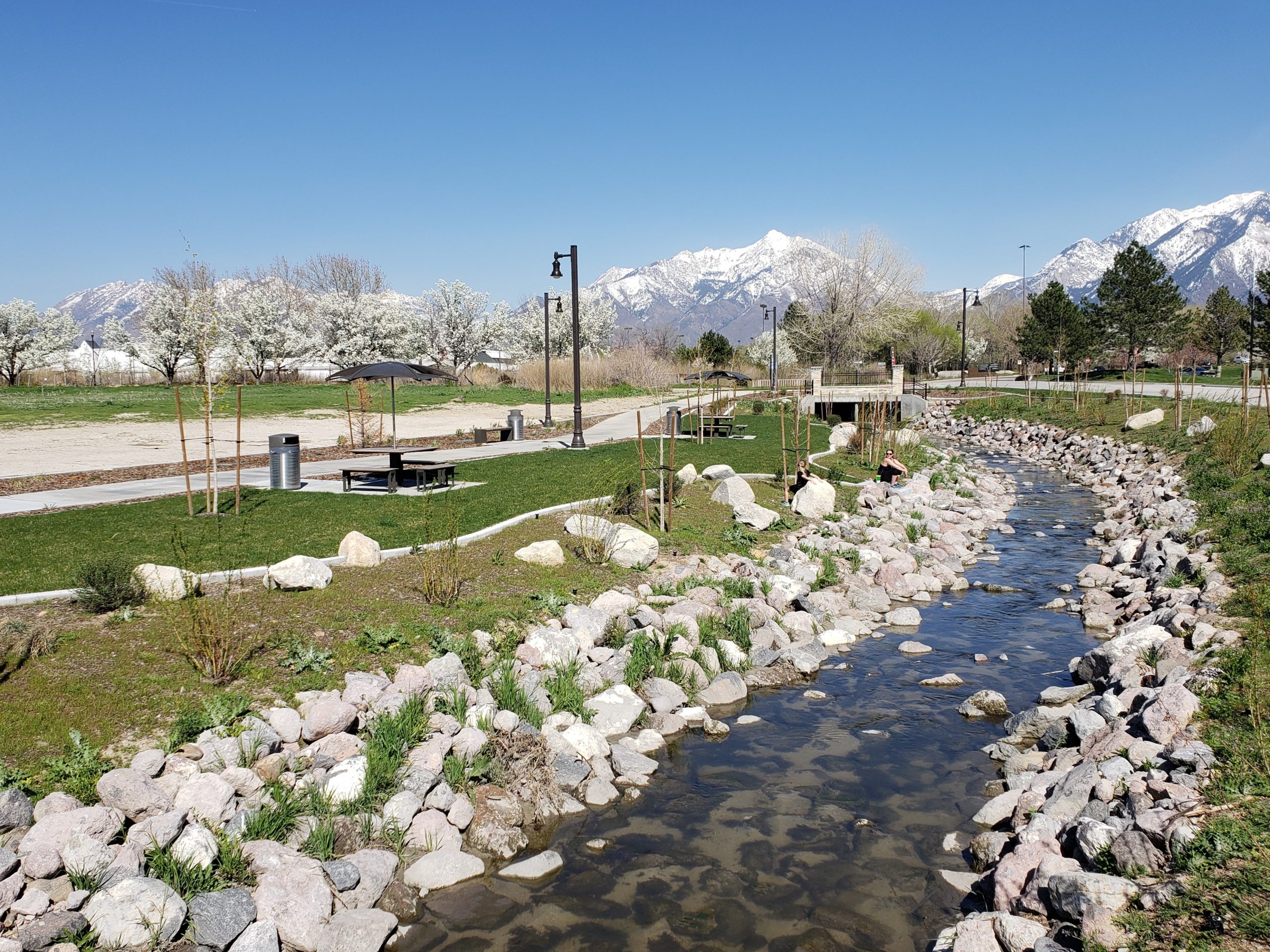 Two people sit beside the Dry Creek channel with sidewalks, tables, grass, blossoming trees, and mountains in the background.