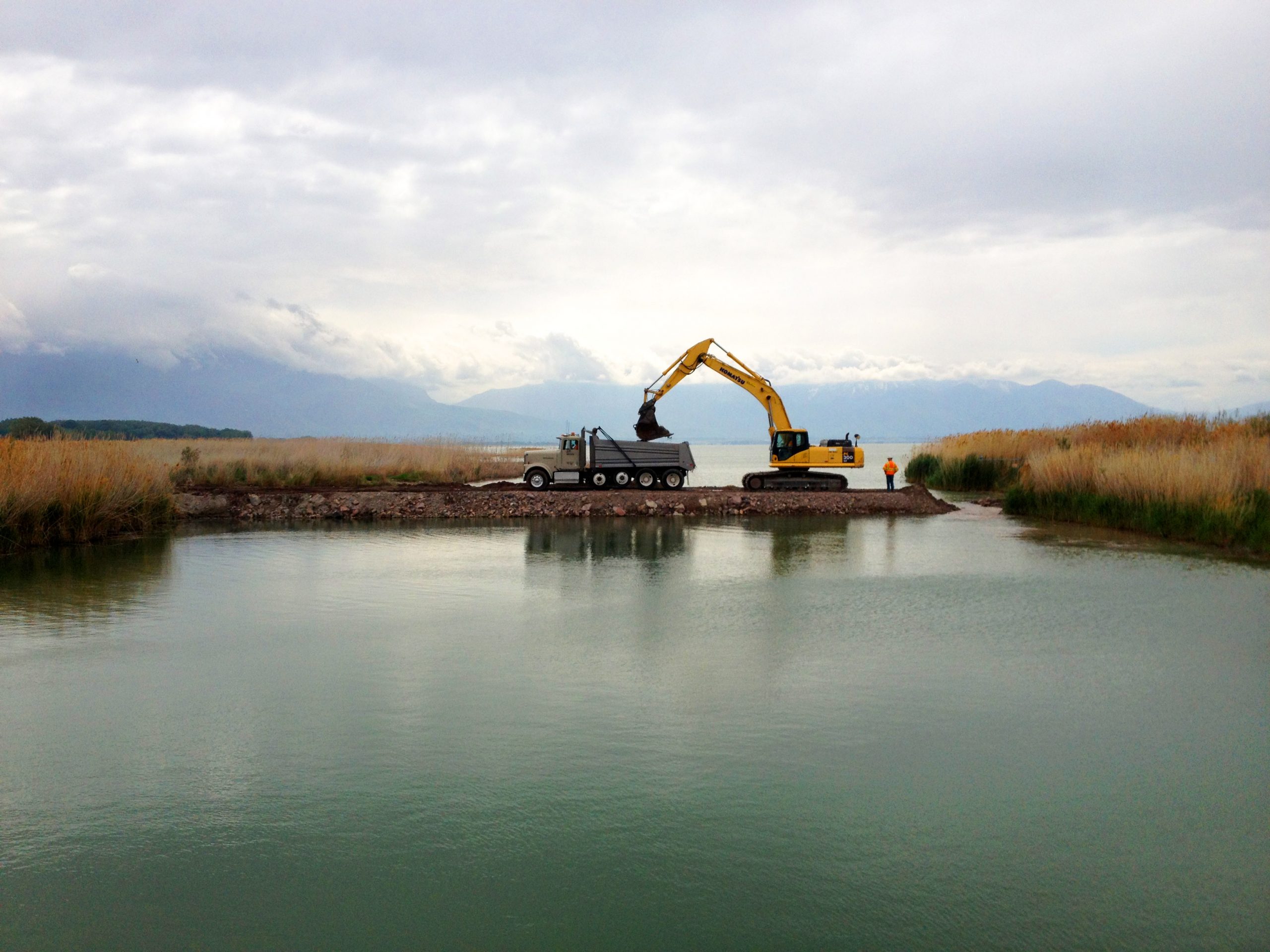 Truck parked at Utah lake as excavator fills tuck bed with dirt.