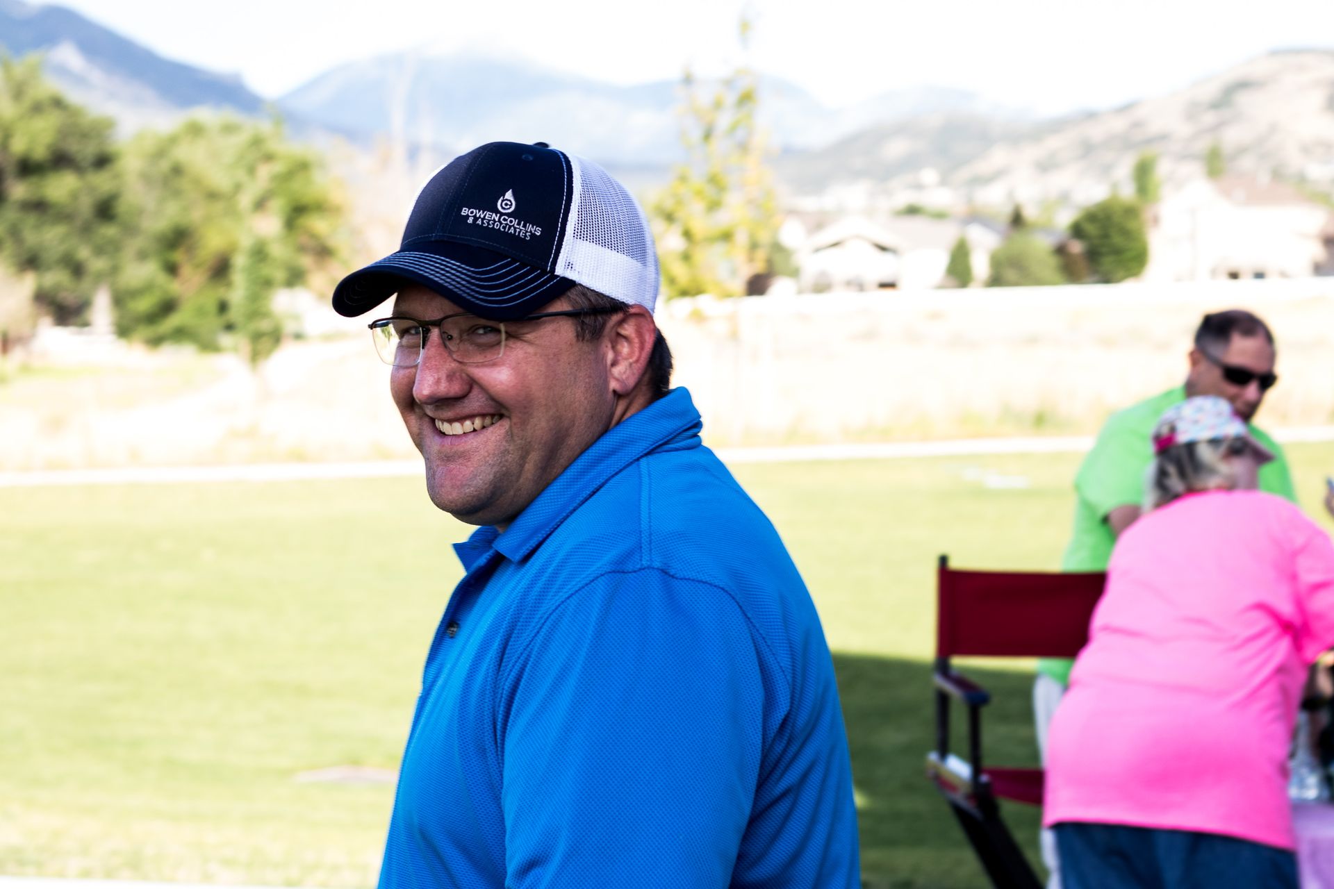Man in blue polo shirt, baseball cap, and glasses smiling.