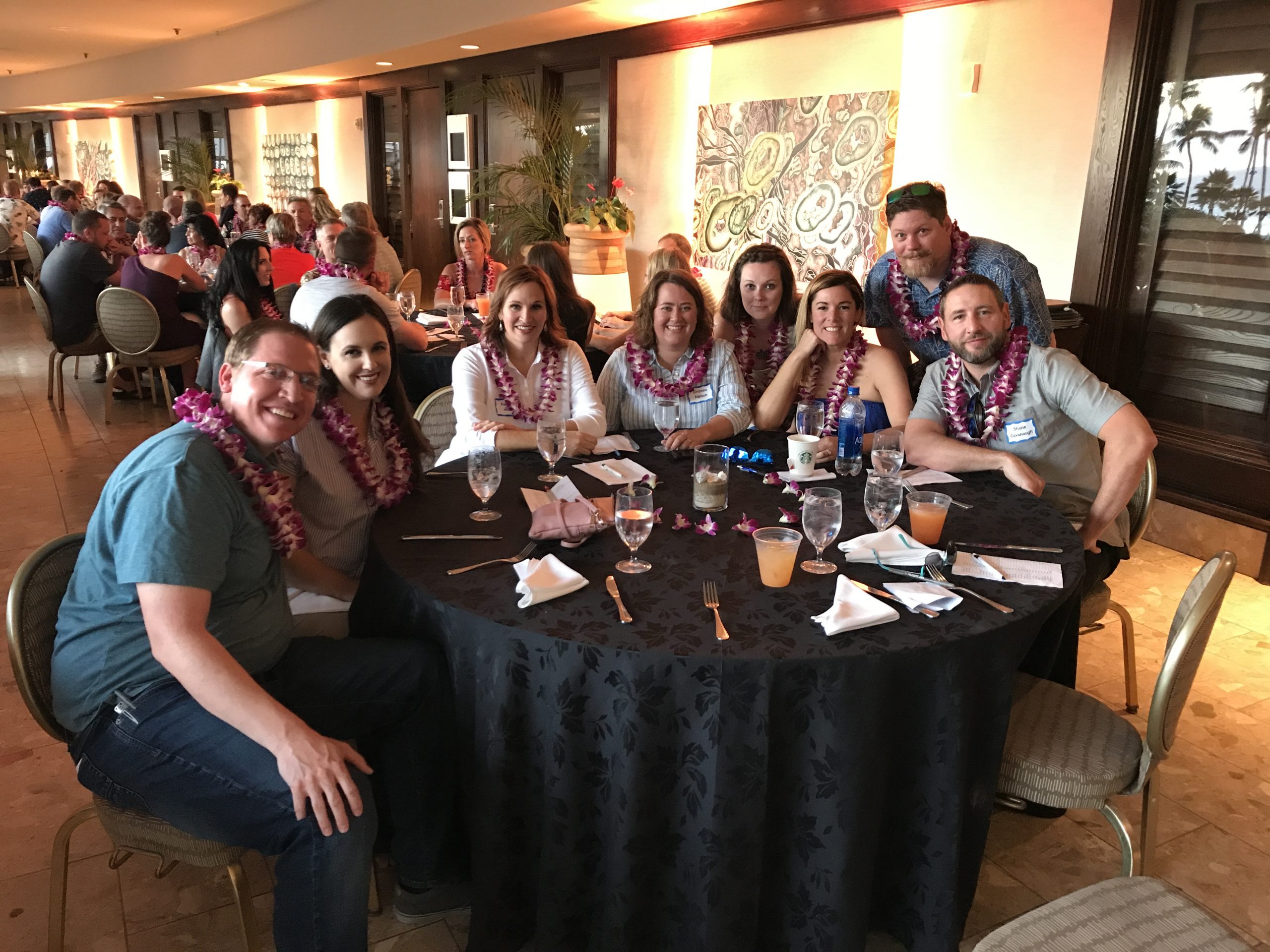 Men and women sitting around dinner table and smiling for photo while wearing leis