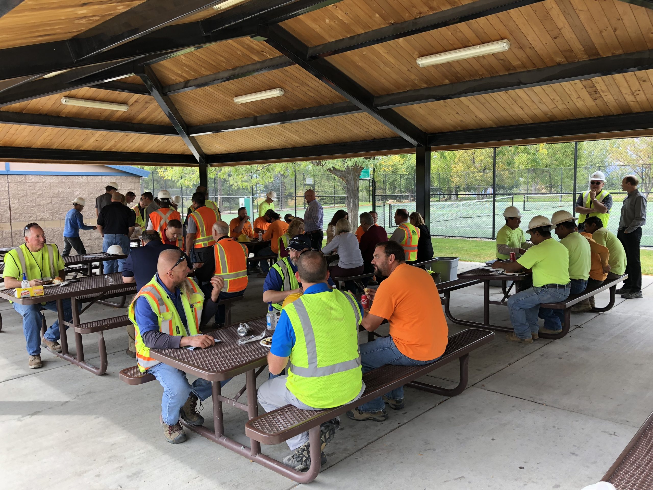 Construction workers sit, talk and eat at metal picnic tables underneath pavilion.